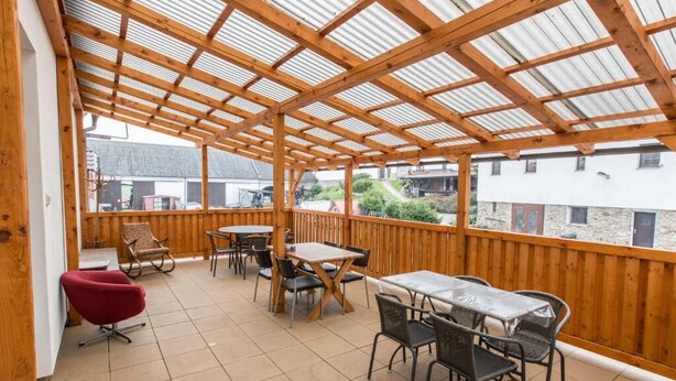 Een overdekt terras buiten, met houten balken en betegelde vloer, met tafels en stoelen.