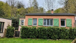 Uma casa térrea com fachada cor de pêssego, persianas verdes e um conjunto de arbustos bem cuidados na frente.