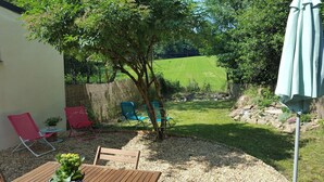 Um pátio com mesa e cadeiras, uma pequena área de jardim com uma árvore, e vista para um campo verde.