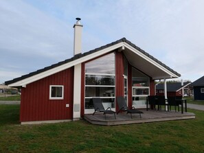 Ein rotes, eingeschossiges Gebäude mit geneigtem Dach, großen Fenstern und einer überdachten Terrasse mit Stühlen.