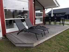 Eine Terrasse mit zwei Liegestühlen und einem Tisch, ein Glastür-Eingang und ein Blick auf ein Haus im Hintergrund.
