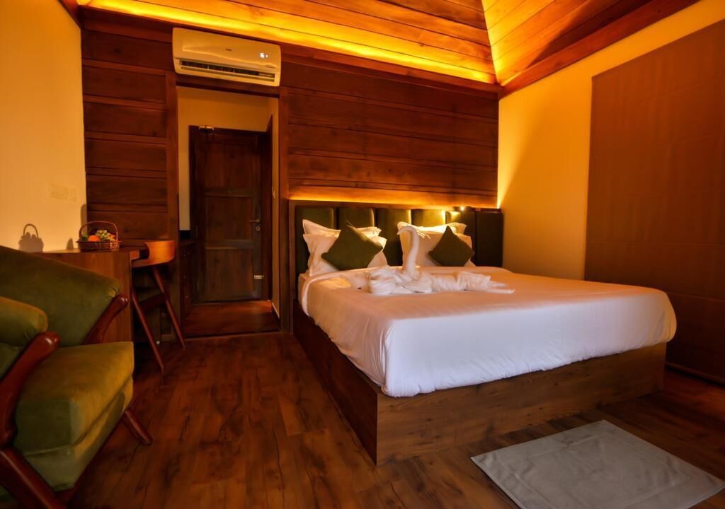 A hotel room with a wooden bed, a green chair, a small table, and an air conditioning unit.