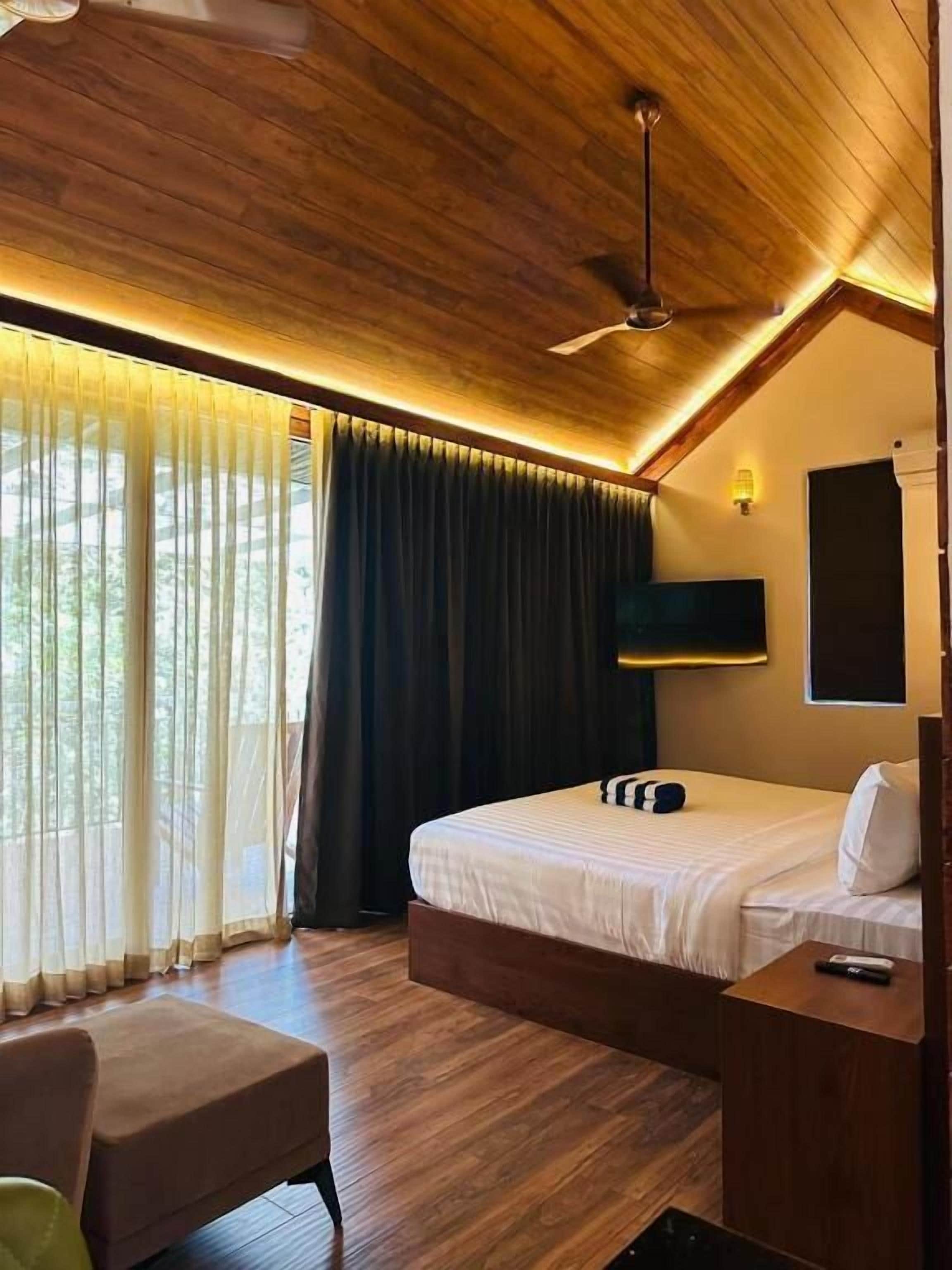 A bedroom with a wooden ceiling, a bed with white bedding, a bedside table, a chair, and a ceiling fan.