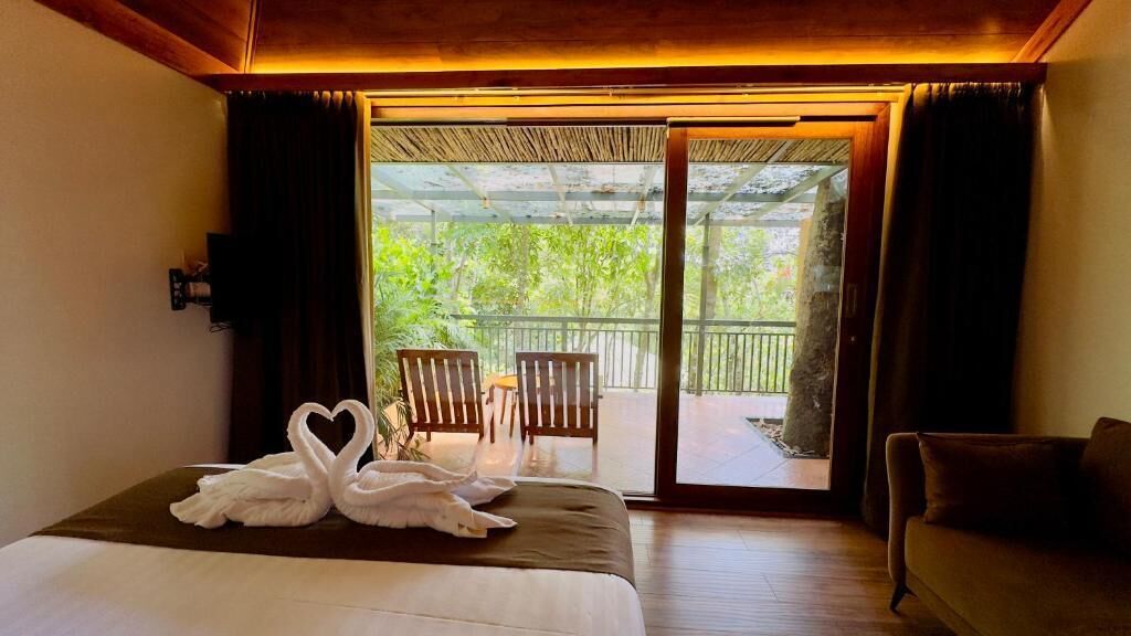 A hotel room with a bed, a sofa, a balcony with chairs, and a view of greenery.