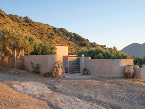 A gravel driveway leading to a gated entrance with a concrete wall, surrounded by vegetation and hills in the background.
