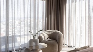 A modern hotel room with a large window, a round table with a vase, a comfortable armchair, and a view of the cityscape. - Moncasa Suites (Agios Nikolaos)