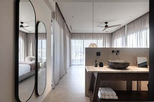 A modern hotel room with a large mirror, a wooden vanity with a stone basin, a ceiling fan, and a view of the city through large windows.