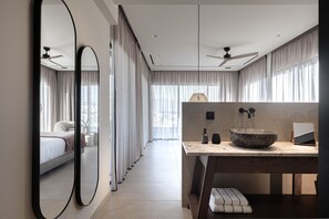 Une chambre d’hôtel moderne dotée d’un grand miroir, d’une coiffeuse en bois avec un lavabo en pierre, d’un ventilateur de plafond et offrant une vue sur la ville grâce à de grandes fenêtres.