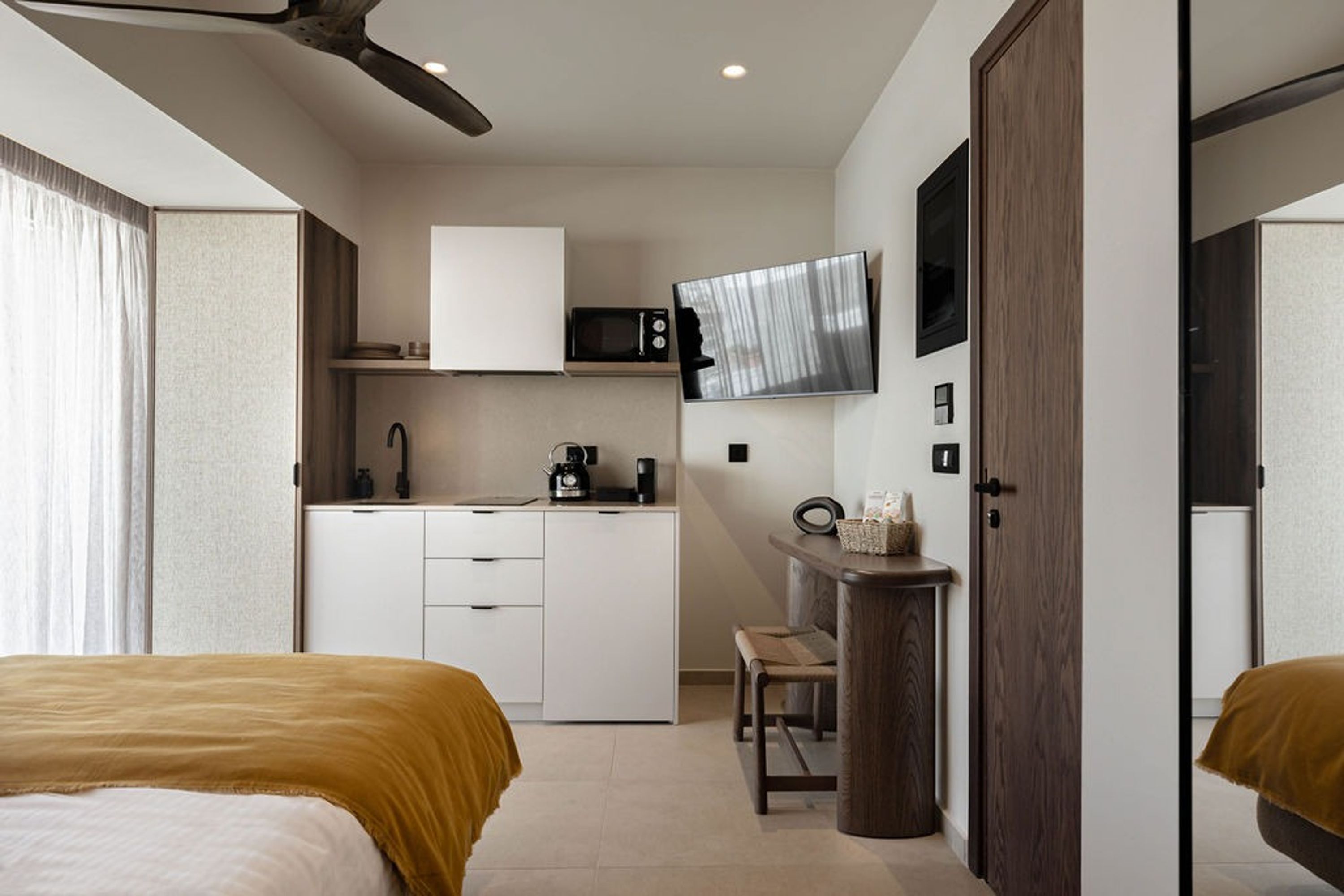 Quarto de hotel pequeno com cozinha compacta, uma cama, uma cadeira e uma TV fixada na parede.