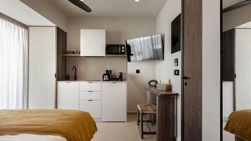 Quarto de hotel pequeno com cozinha compacta, uma cama, uma cadeira e uma TV fixada na parede.