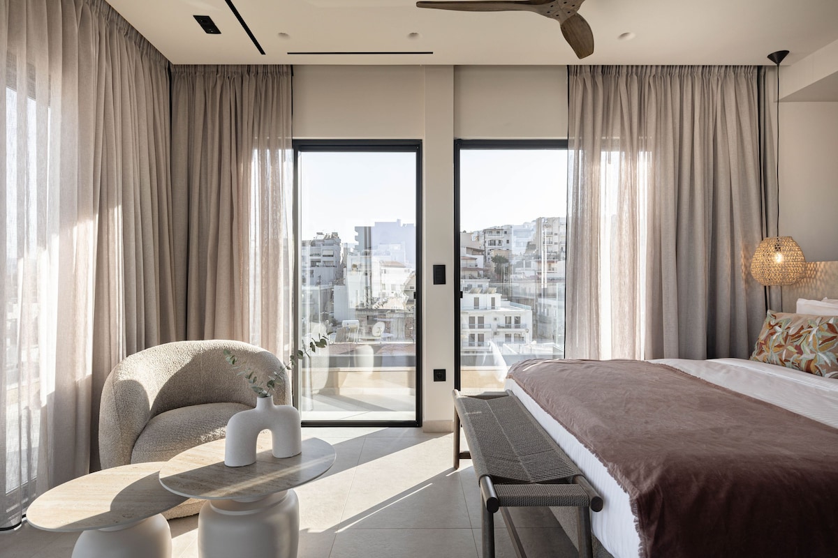 A modern hotel room with a bed, a seating area, a table, and a view of the cityscape through large windows.