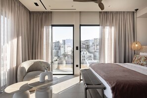 A modern hotel room with a bed, a seating area, a table, and a view of the cityscape through large windows. - Moncasa Suites (Agios Nikolaos)