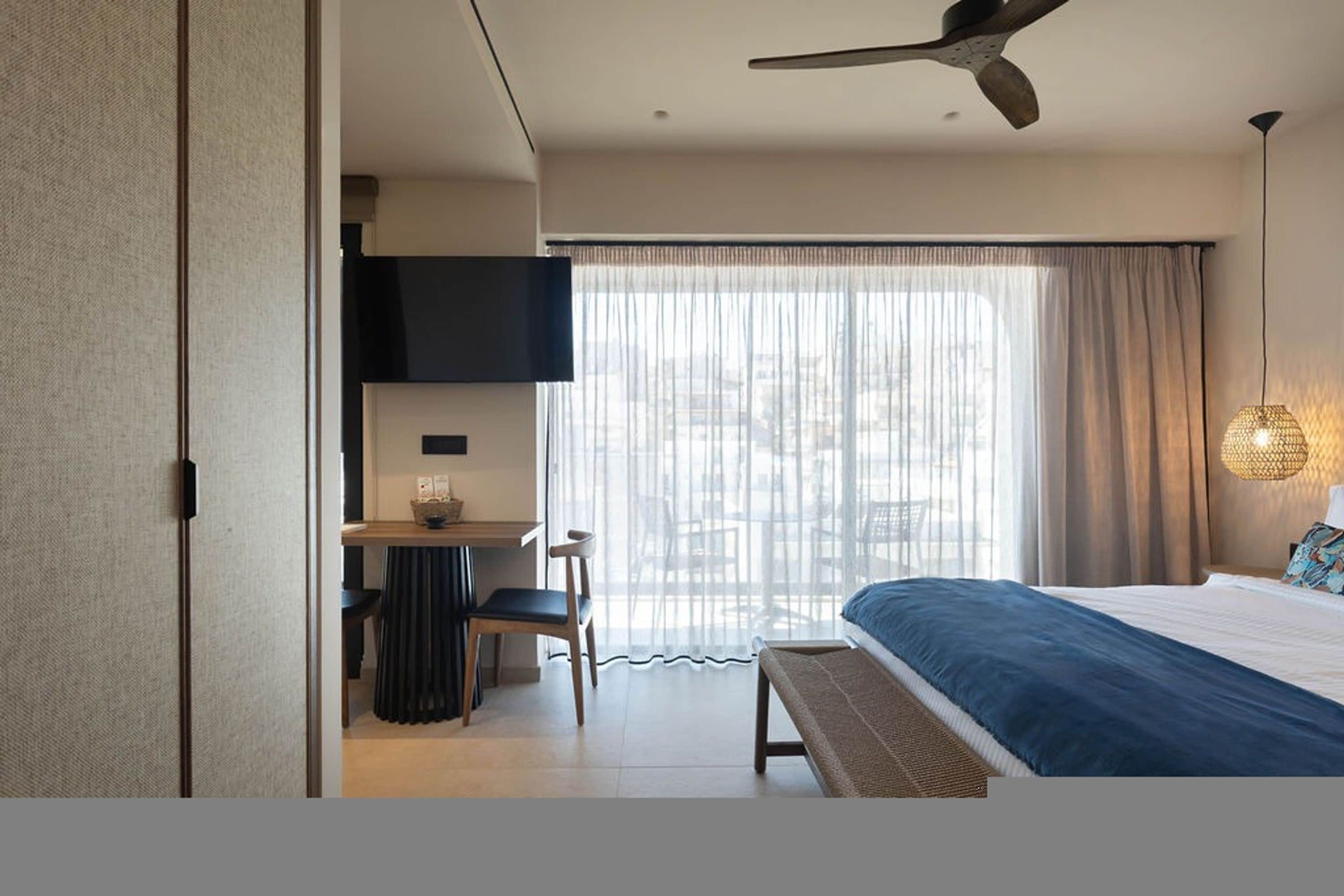 A modern hotel room with a bed, a desk, a chair, a television, and a large window with curtains.