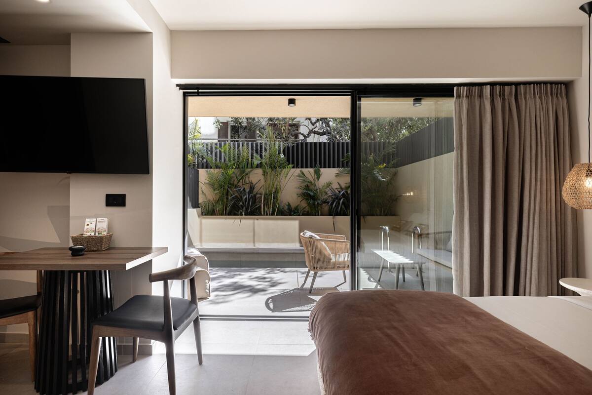 A modern hotel room with a large bed, a dining area, a TV, and a balcony with plants.