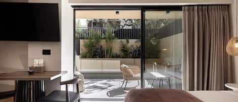 A modern hotel room with a large bed, a dining area, a TV, and a balcony with plants.