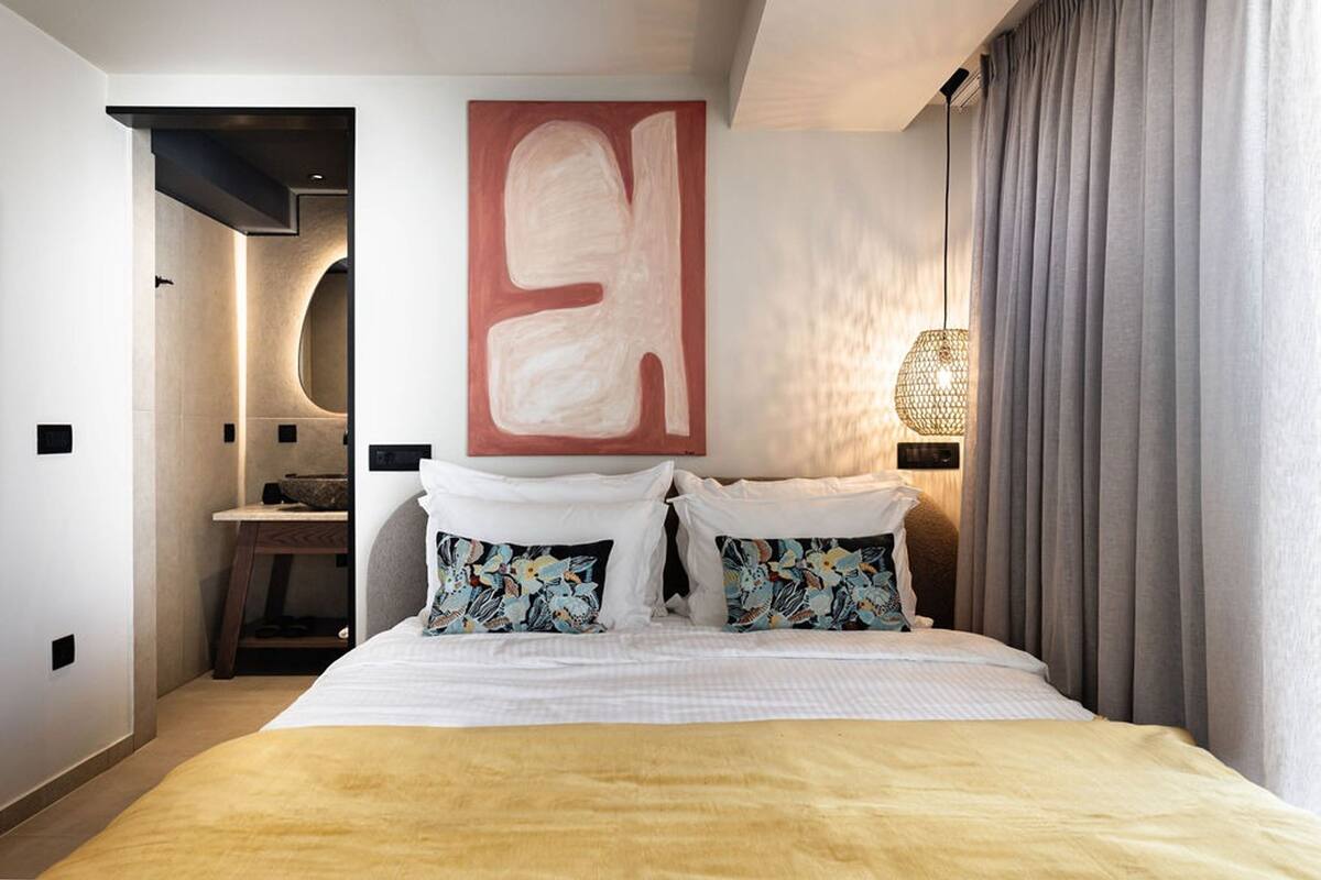 A modern hotel room with a large bed, a wall-mounted abstract painting, a bedside lamp, and a view of a cityscape through the window.