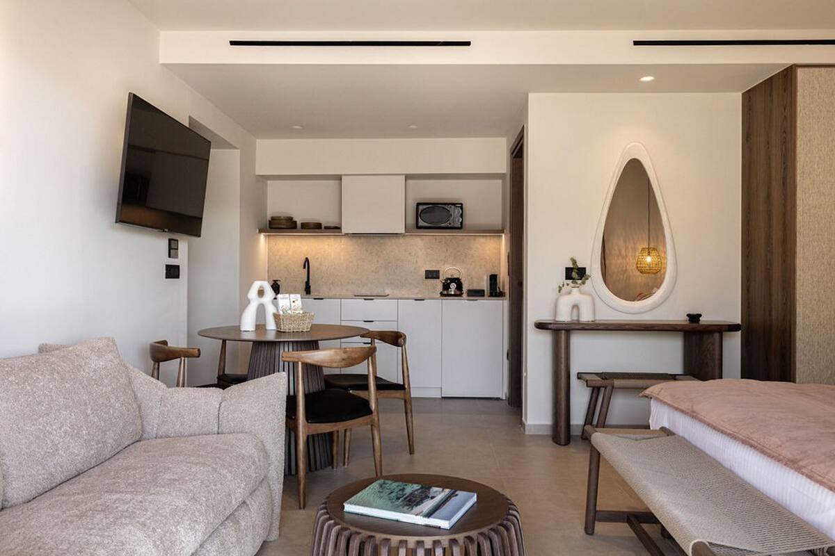 A modern hotel room with a sofa, a small round table with a book, a bench, a TV mounted on the wall, a kitchenette, and a large mirror.