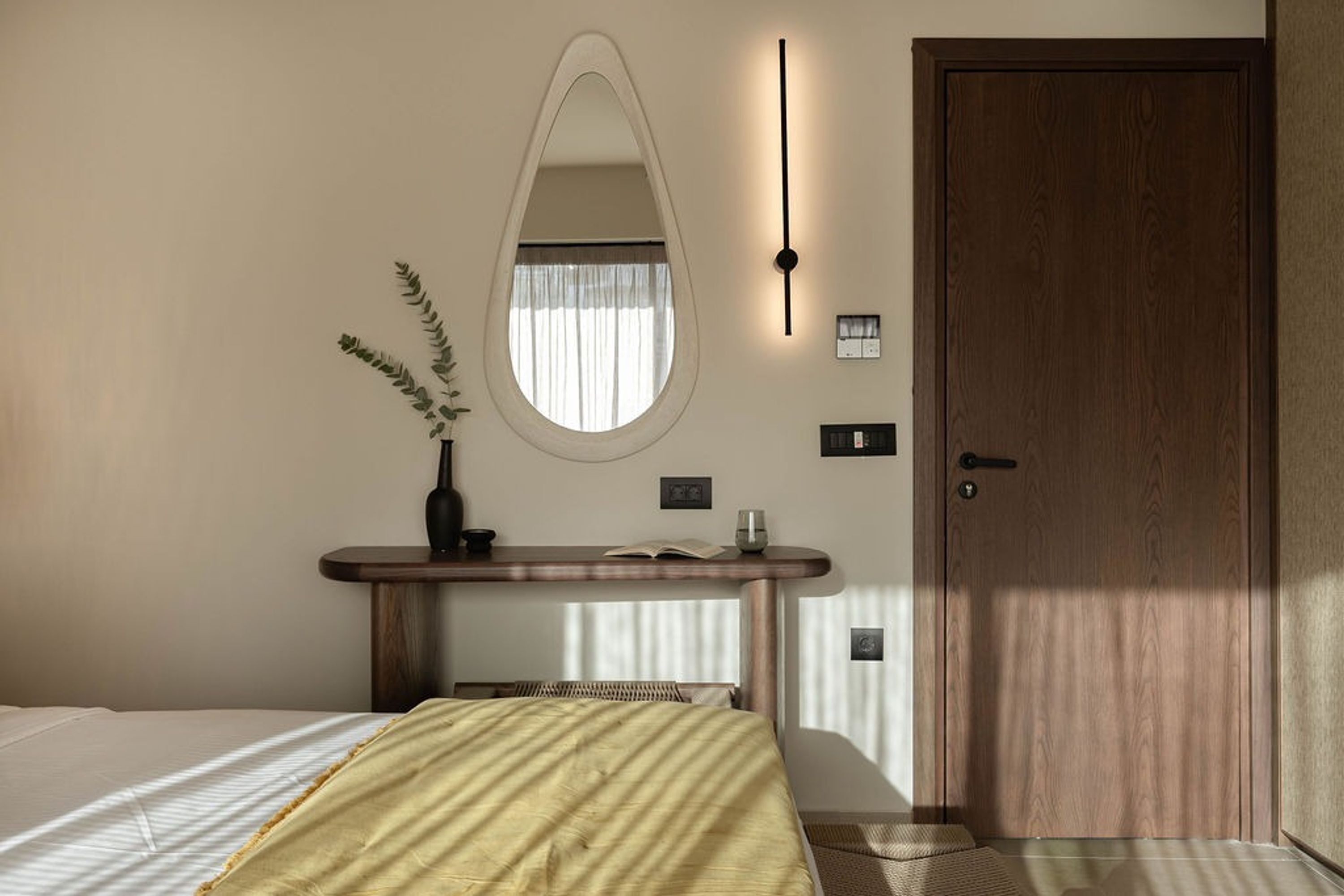 A bedroom with a bed, a bedside table with a vase, a mirror, a door, and a wall-mounted light.