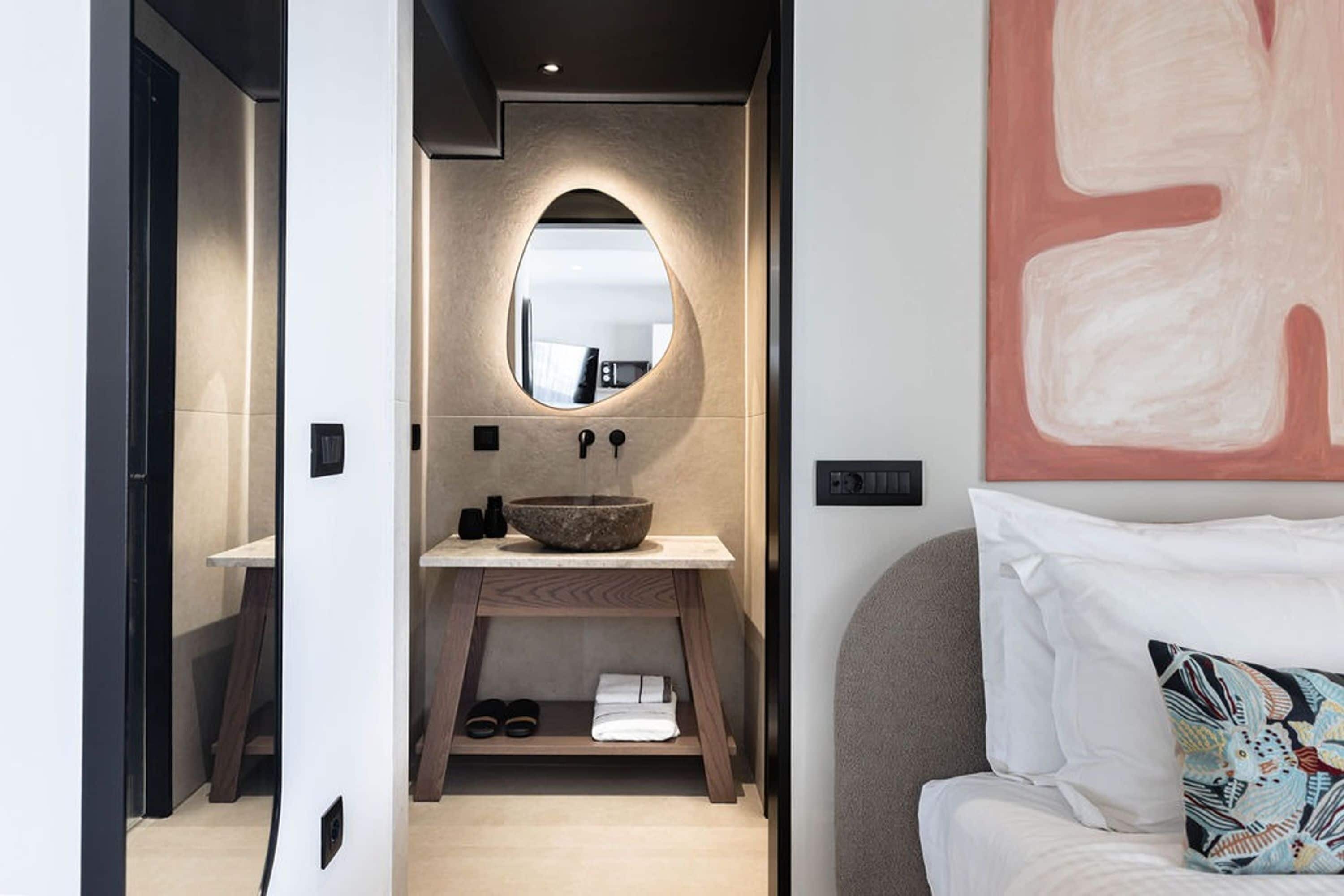 A modern bathroom with a large oval mirror, a stone sink, and a wooden shelf with slippers.