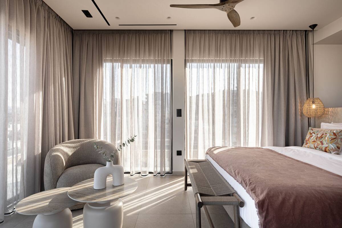 A modern hotel room with a large bed, a seating area, and a view through sheer curtains.