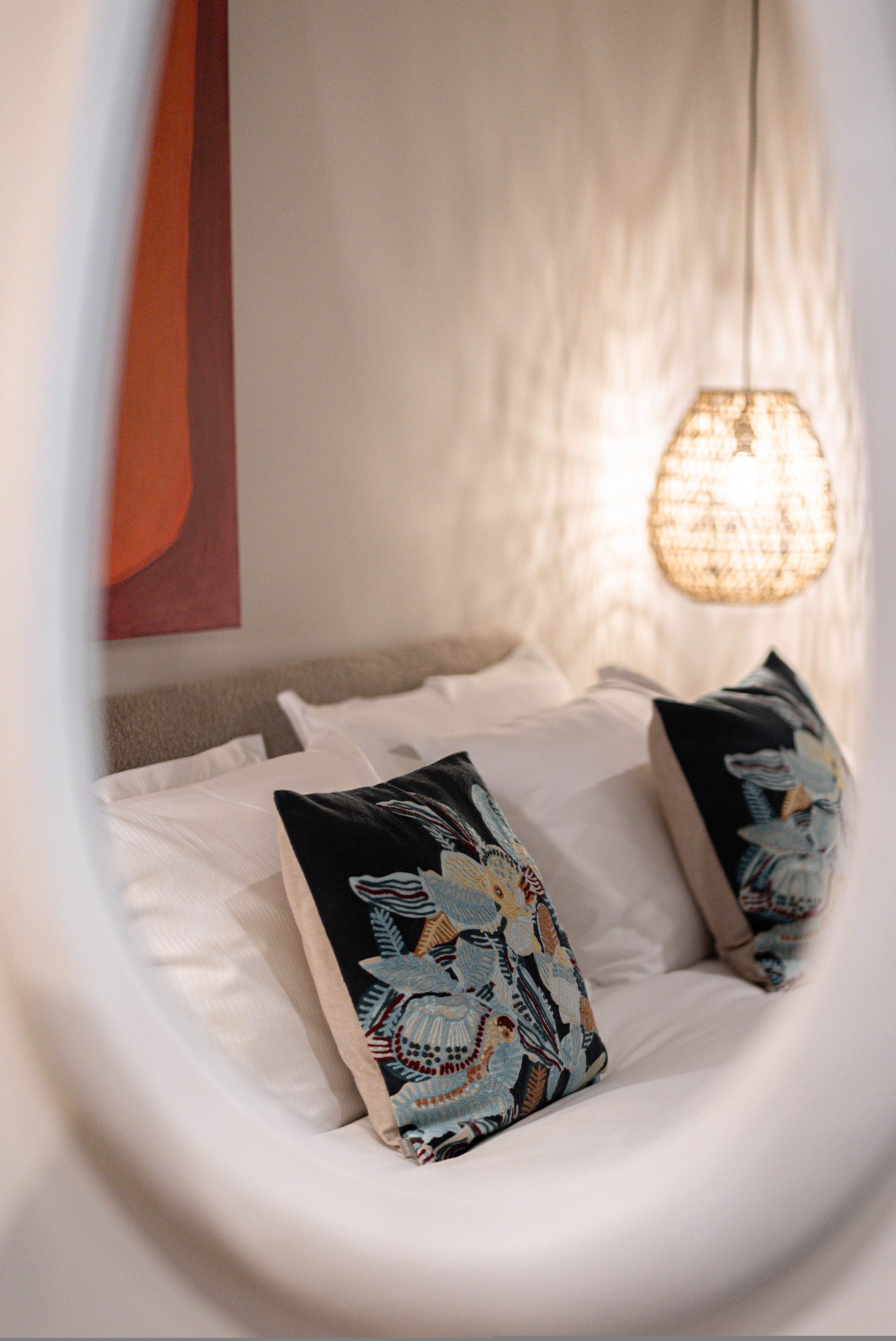 Ein Bett mit weißer Bettwäsche und gemusterten Kissen, eine Hängeleuchte und ein Teil einer Wand mit rotem Akzent.