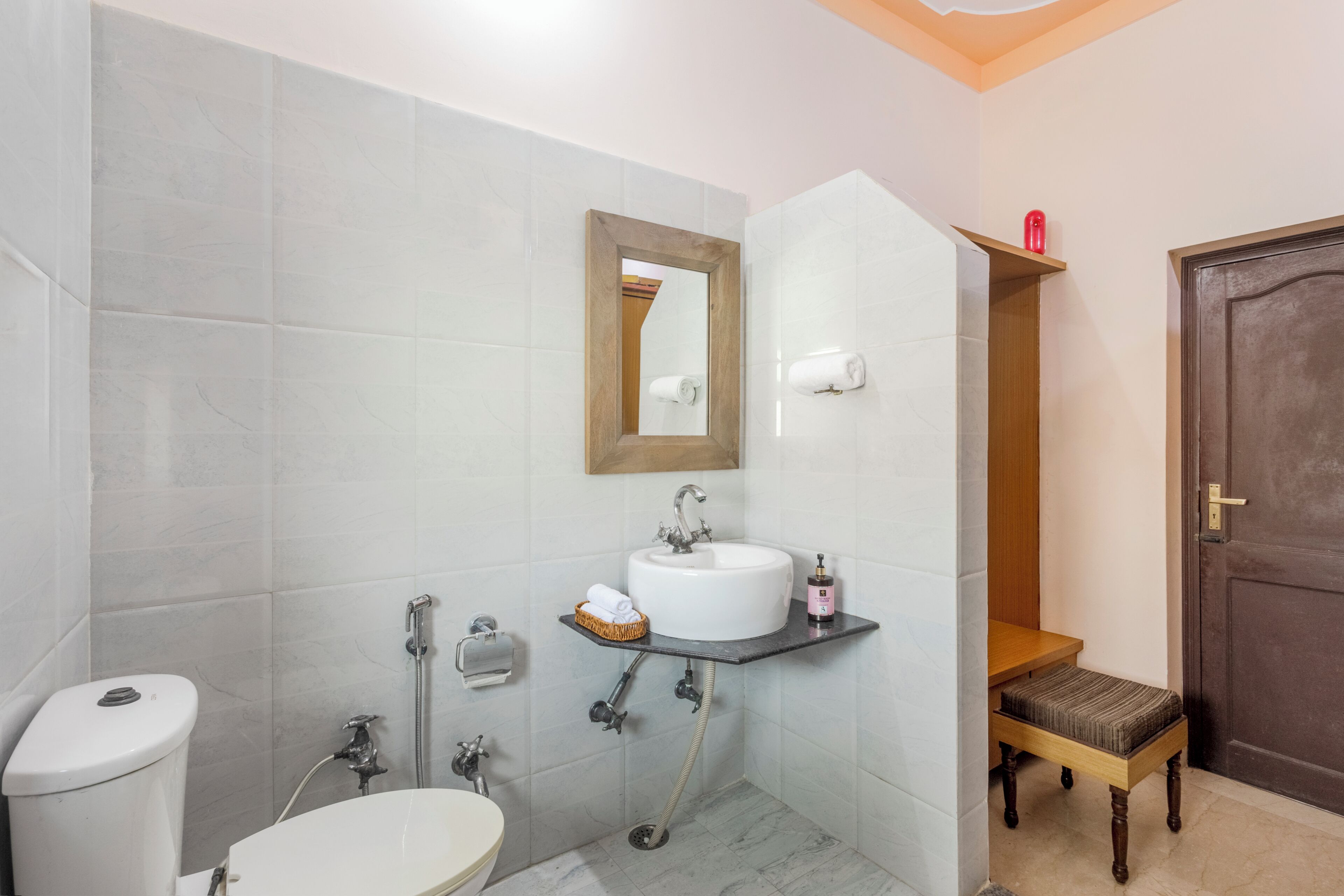 Un baño con inodoro, lavamanos, espejo, toallero y un banco de madera.