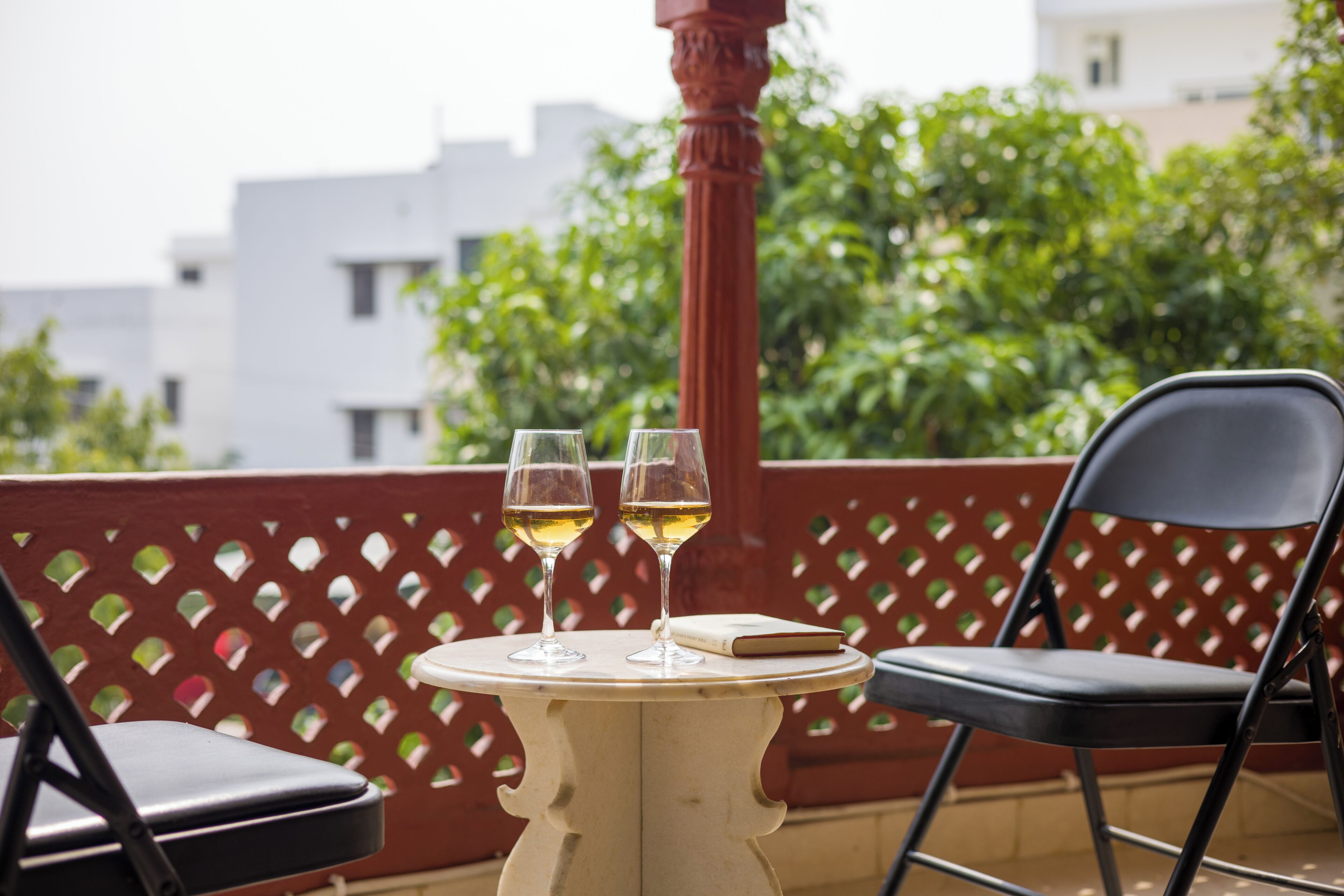 A round table with two glasses of wine, a book, and a black chair on a balcony with a lattice railing and greenery in the background.