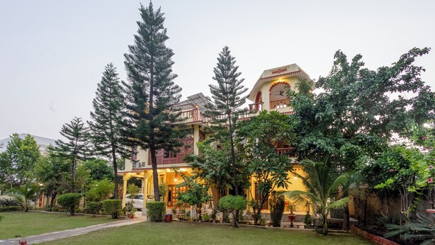A large building with multiple floors, a clock tower, and a well-maintained garden with various trees and shrubs. - Alaya Stays 4 BHK with Outdoor Dining Area - Abhimanyu Mansion Sapphire (Udaipur)