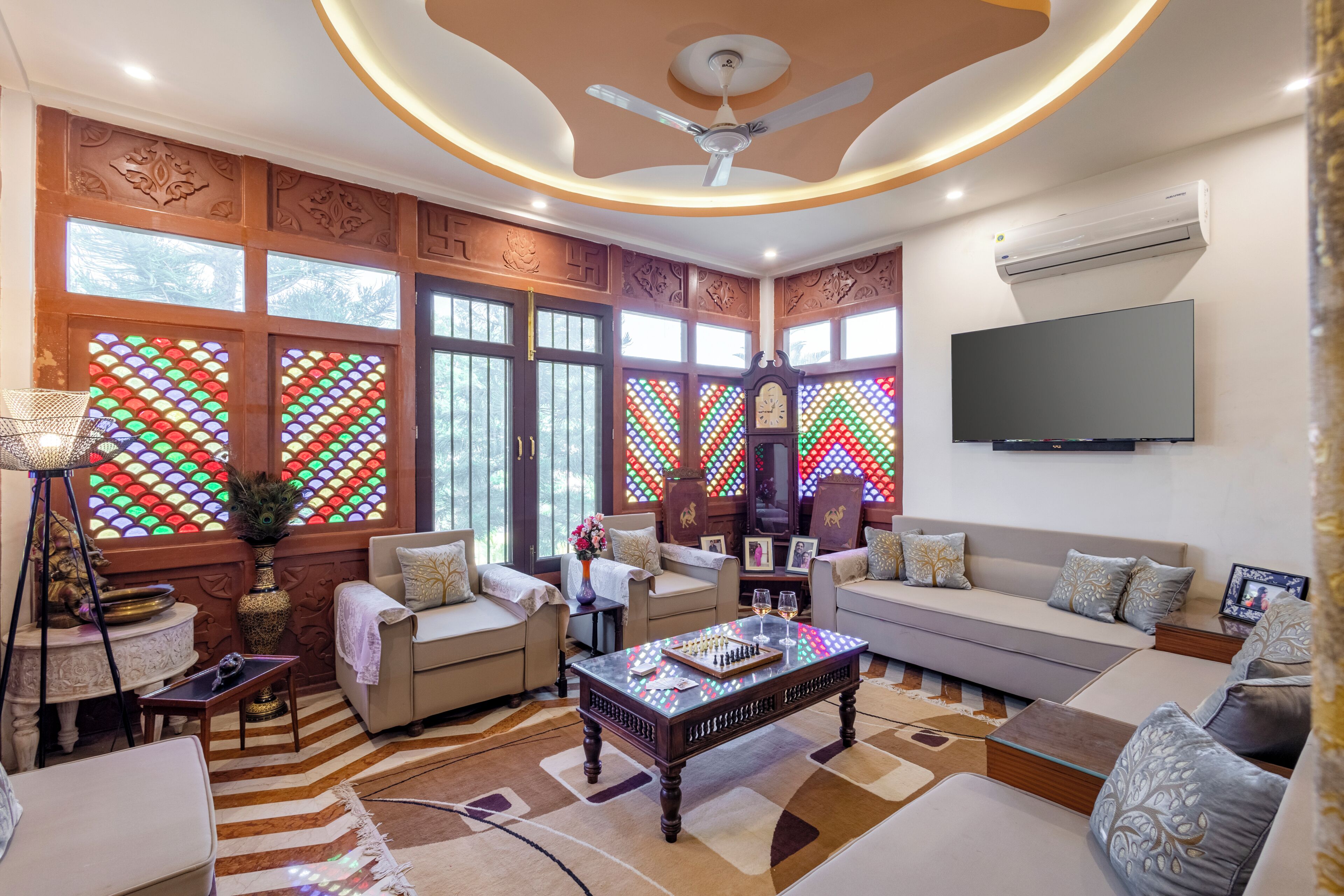 A spacious living room with a ceiling fan, large windows with stained glass, a flat-screen TV, and a sofa set.