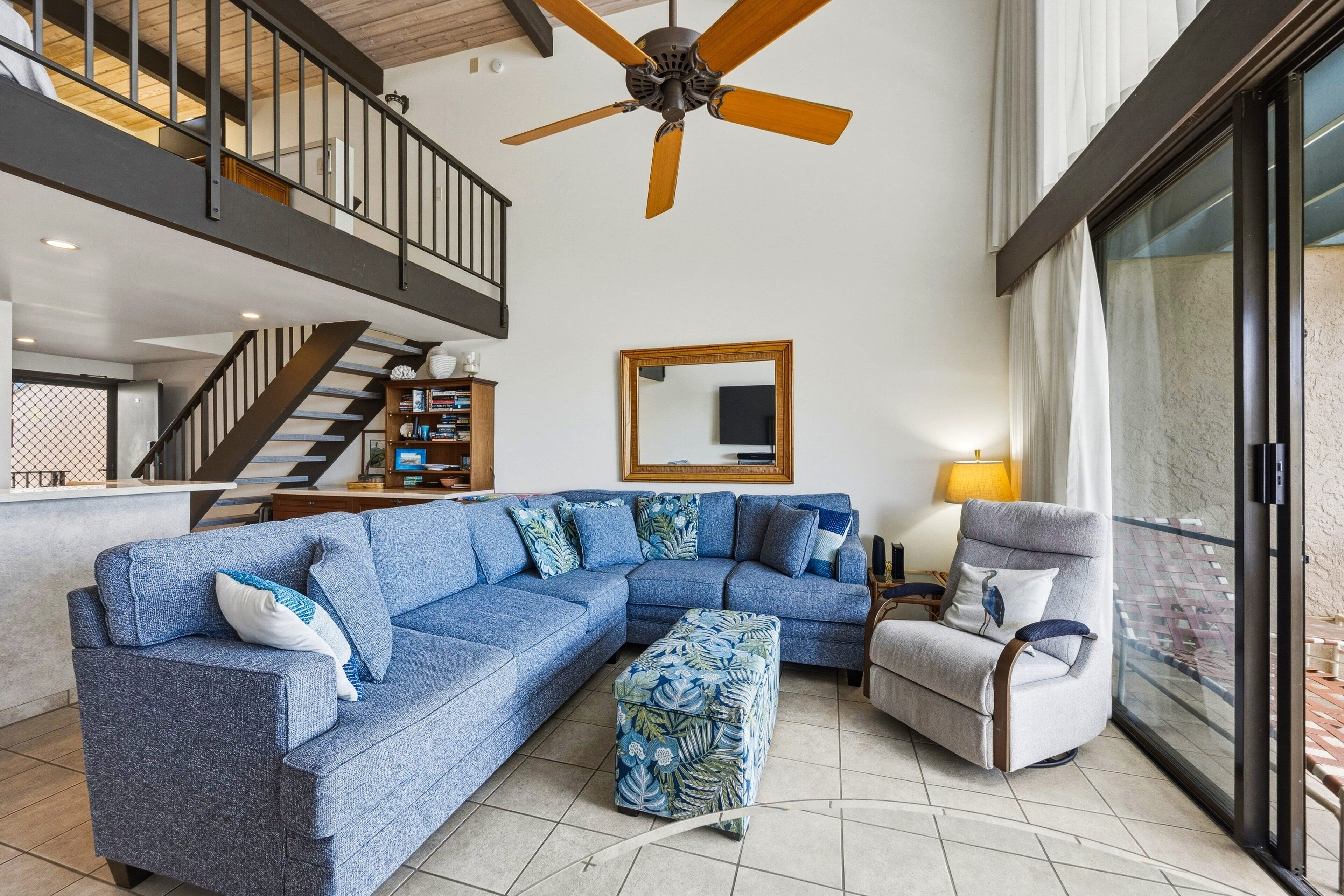 Ein Wohnzimmer mit einem Ecksofa, einer Liege, einem Deckenventilator, einem großen Spiegel und Blick durch eine Glastür.