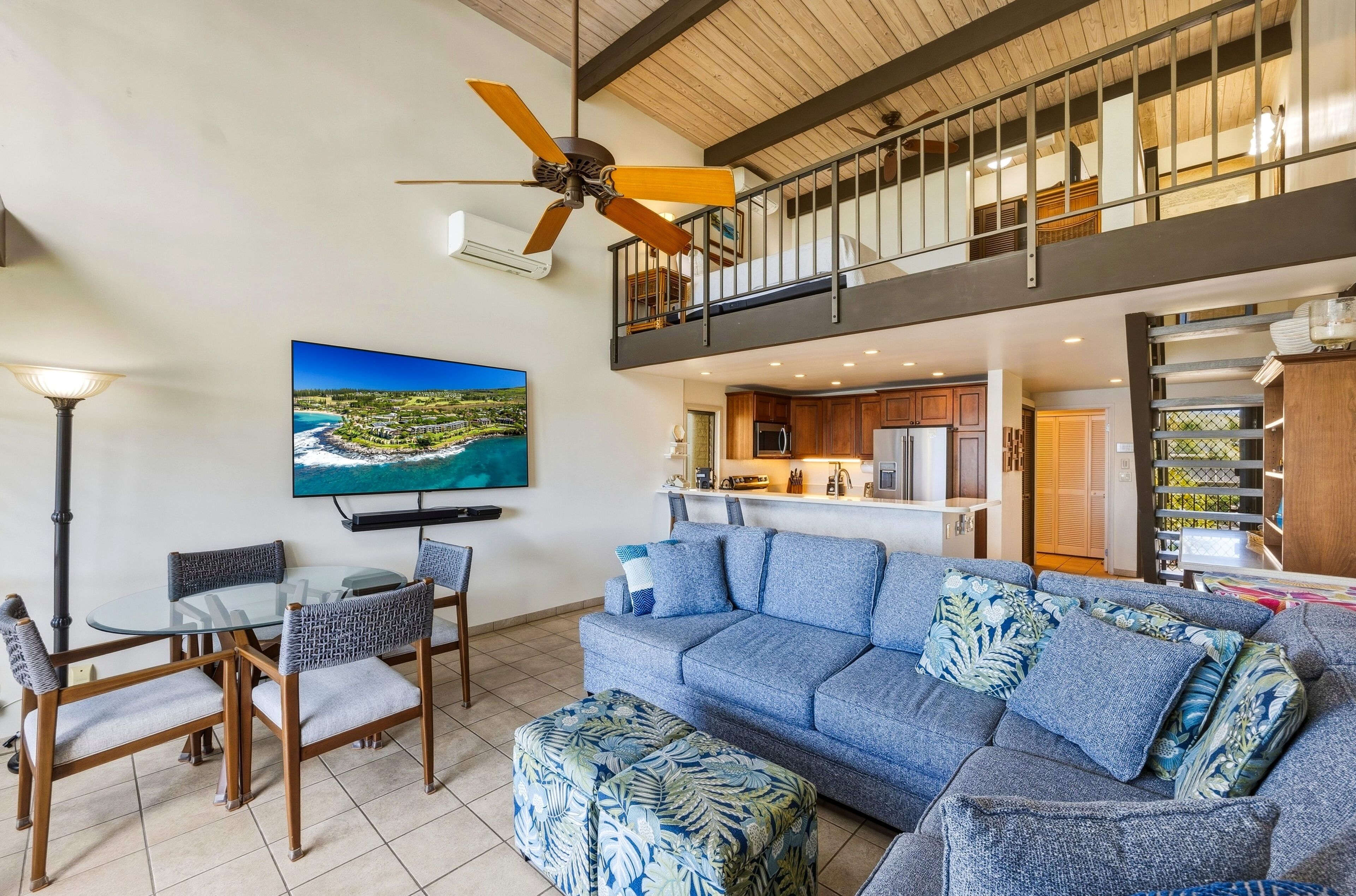 A spacious living area with a sofa, chairs, a dining table, a ceiling fan, a TV, and a kitchen in the background.