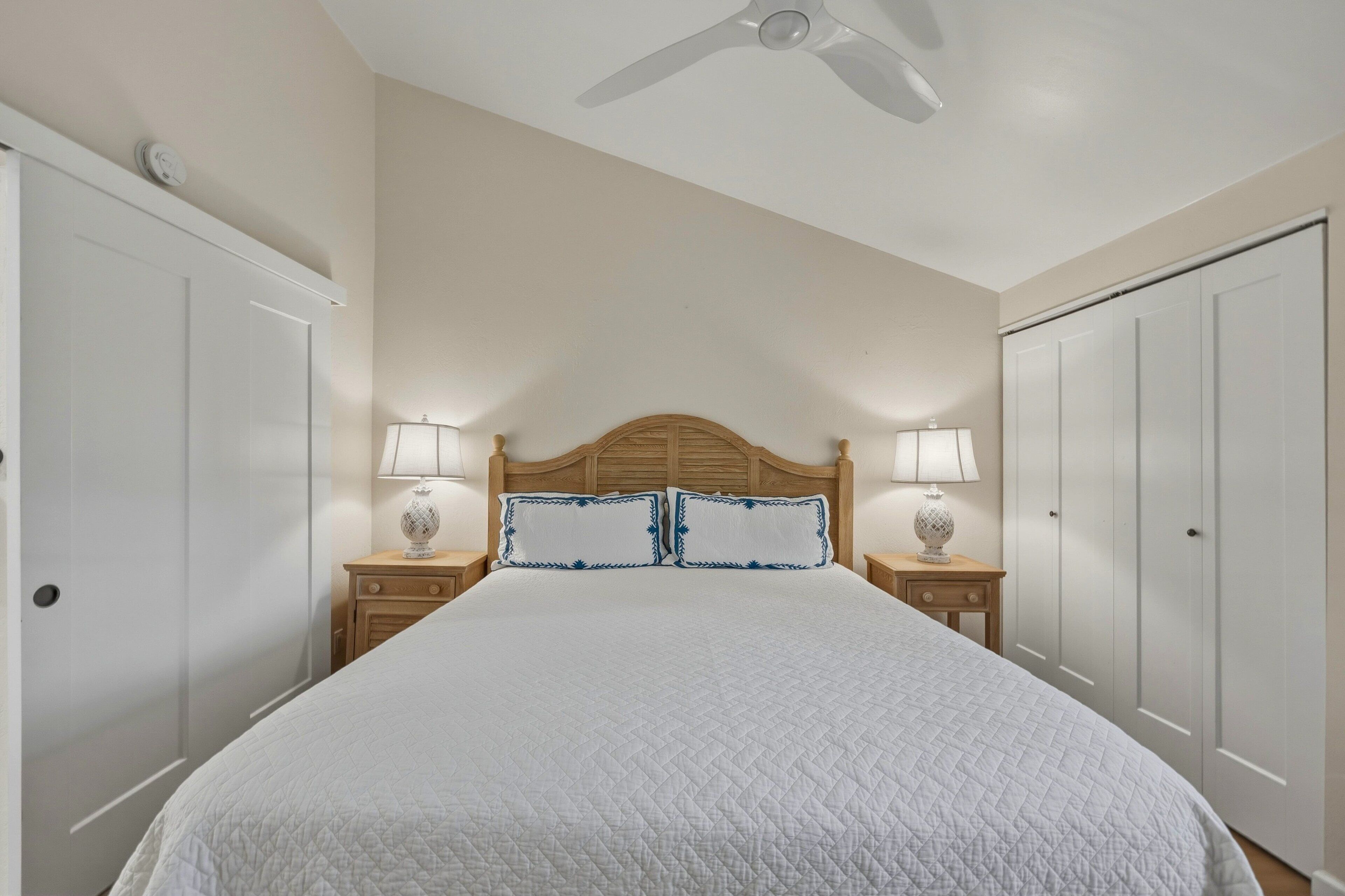 Une chambre avec un grand lit, deux tables de chevet avec des lampes, un ventilateur de plafond et des armoires blanches.