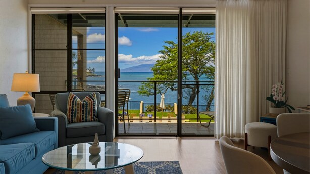 A modern living room with a glass door leading to a balcony with a view of the sea and mountains. - Prime Oceanfront 2br Loft - Unit C10 1 Bedroom Condo (Lahaina)
