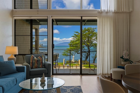 A modern living room with a glass door leading to a balcony with a view of the sea and mountains. - Prime Oceanfront 2br Loft - Unit C10 1 Bedroom Condo (Lahaina)