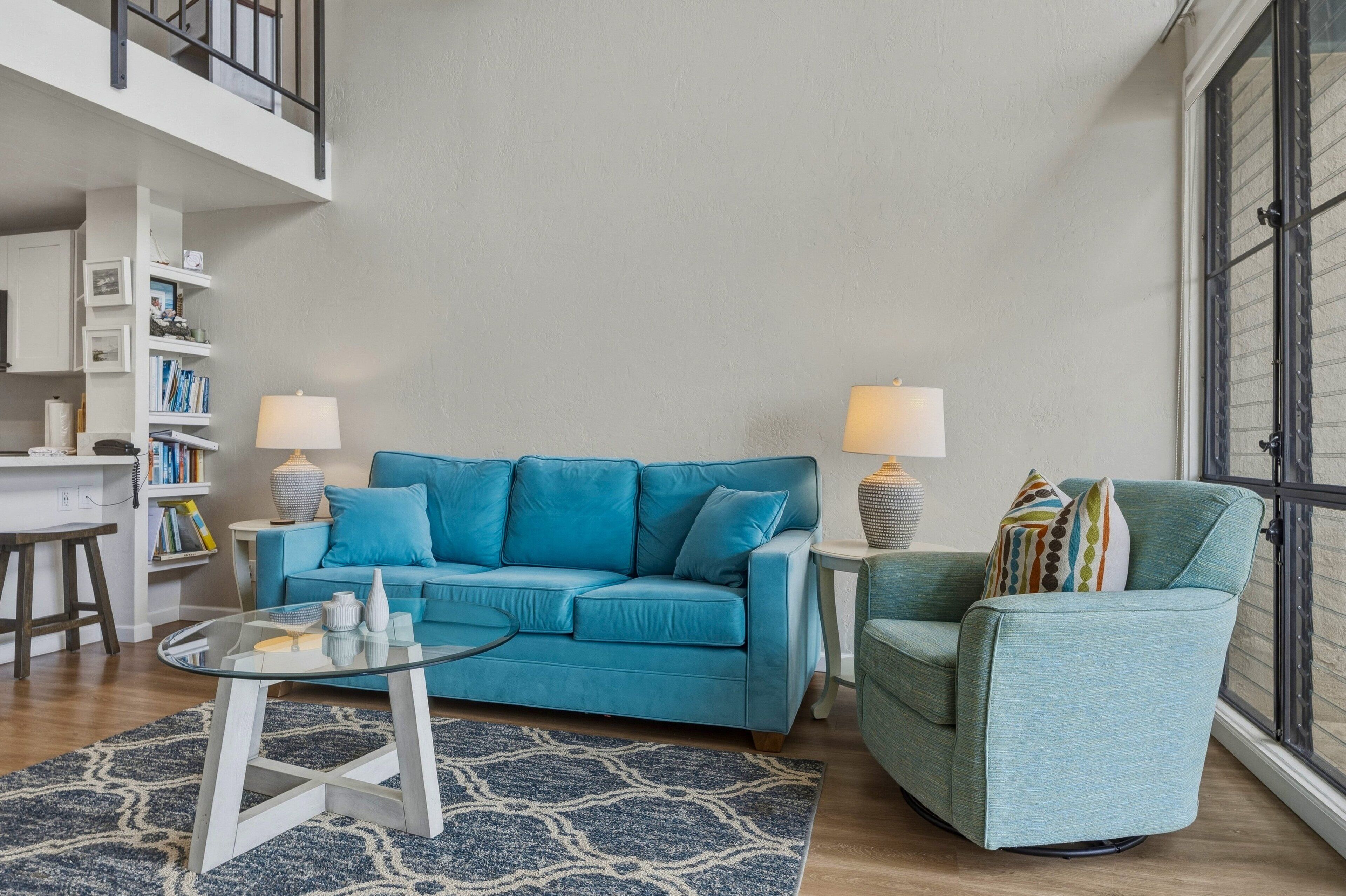 Una sala de estar con un sofá azul, un sillón verde, una mesa de centro de vidrio y dos lámparas.