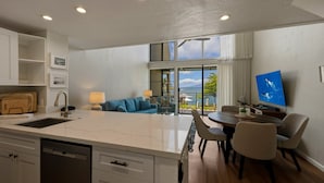 A modern kitchen with a central island, dining area with chairs, a sofa, and a TV. - Prime Oceanfront 2br Loft - Unit C10 1 Bedroom Condo (Lahaina)