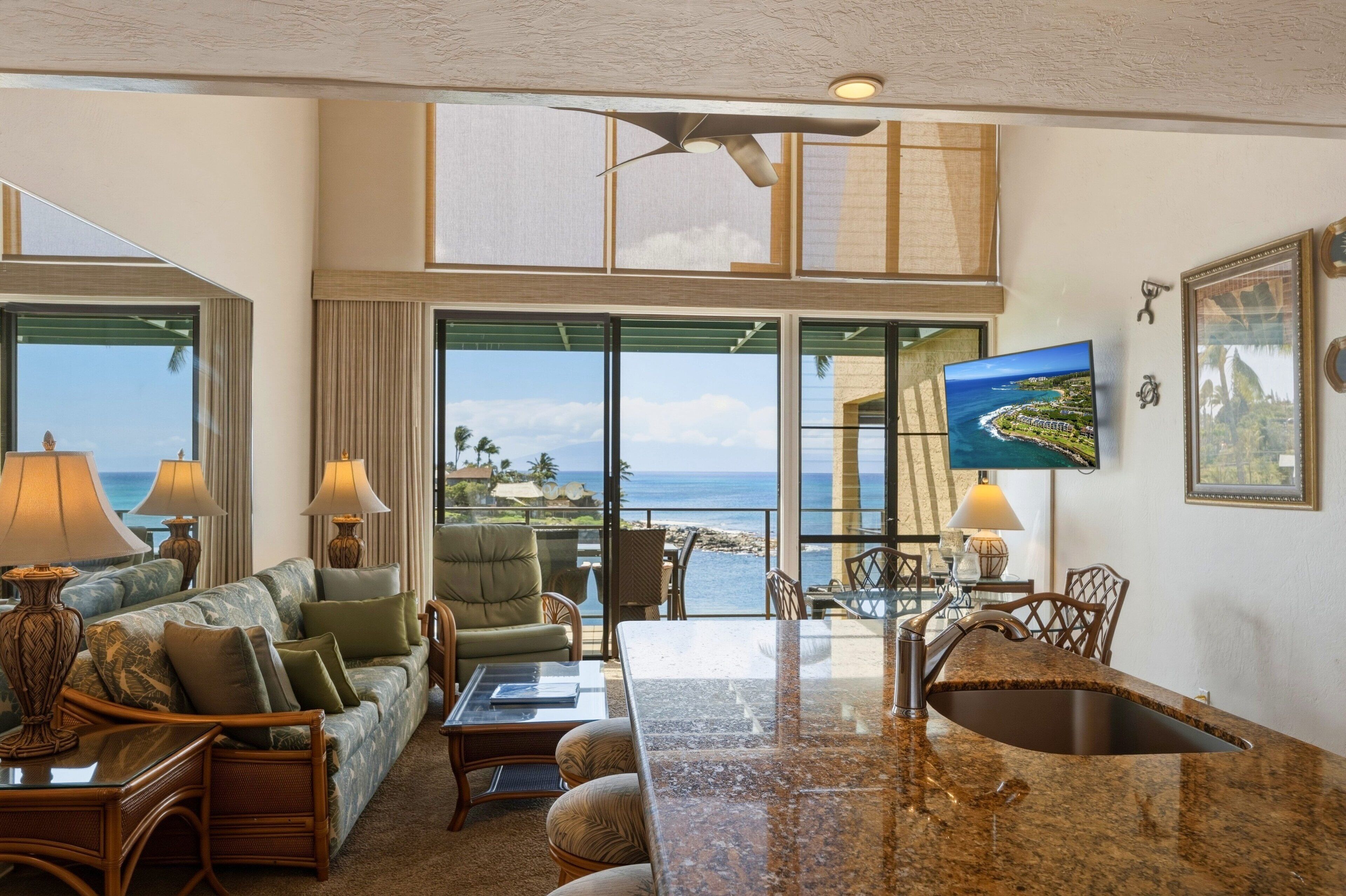 A spacious living area with a large window overlooking the ocean, a ceiling fan, and a flat-screen TV mounted on the wall.