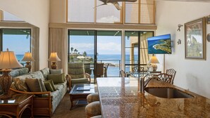 A spacious living area with a large window overlooking the ocean, a ceiling fan, and a flat-screen TV mounted on the wall. - Oceanfront 2br Loft With Sunset Views - Unit C23 1 Bedroom Condo (Lahaina)