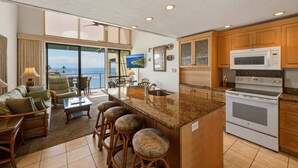 A kitchen with a large island, a microwave, and a stove. There is a dining area with a view of the ocean. - Oceanfront 2br Loft With Sunset Views - Unit C23 1 Bedroom Condo (Lahaina)