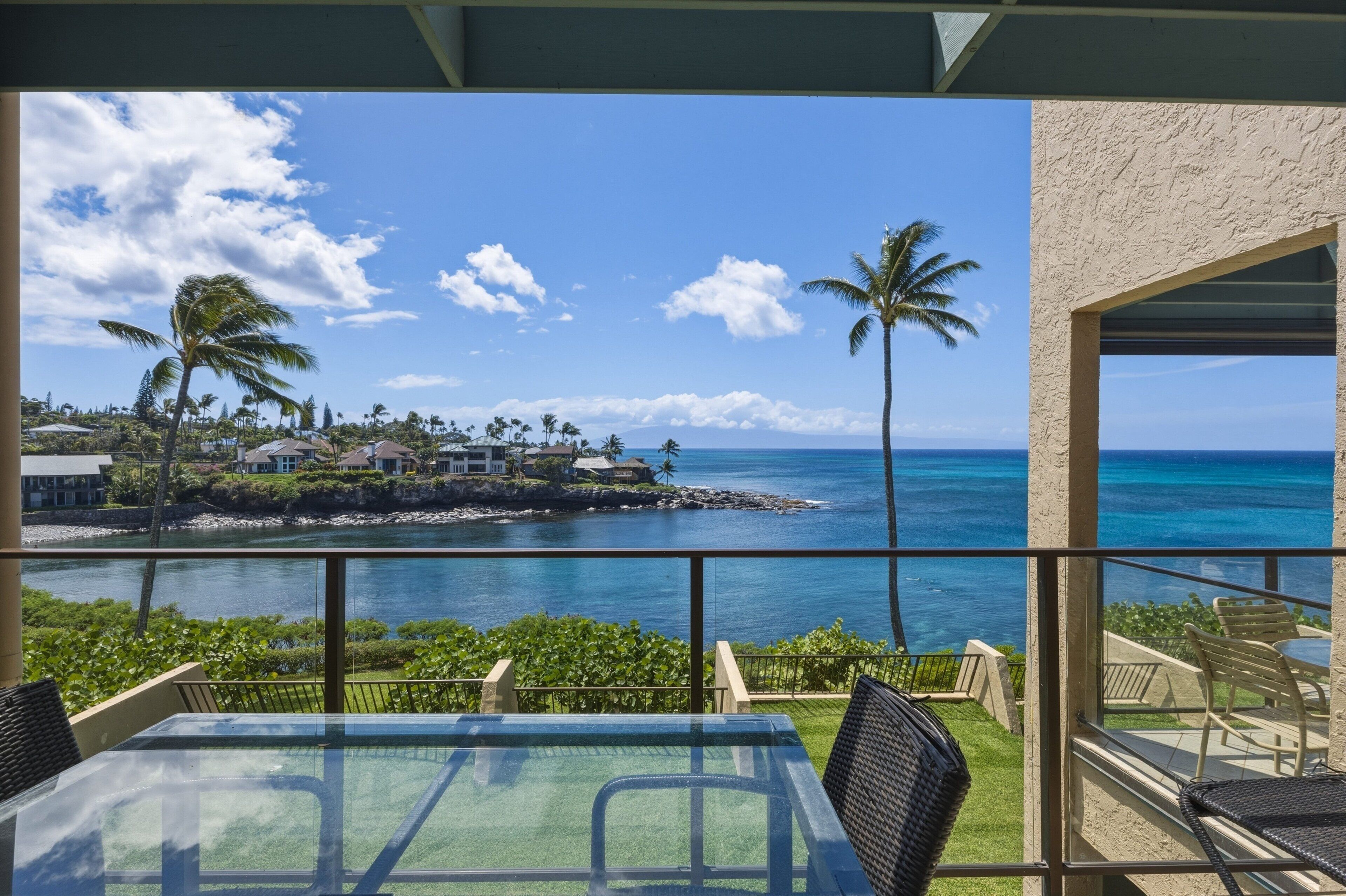 Un balcon avec une table et des chaises en verre, offrant une vue sur la côte, avec des palmiers et des maisons.