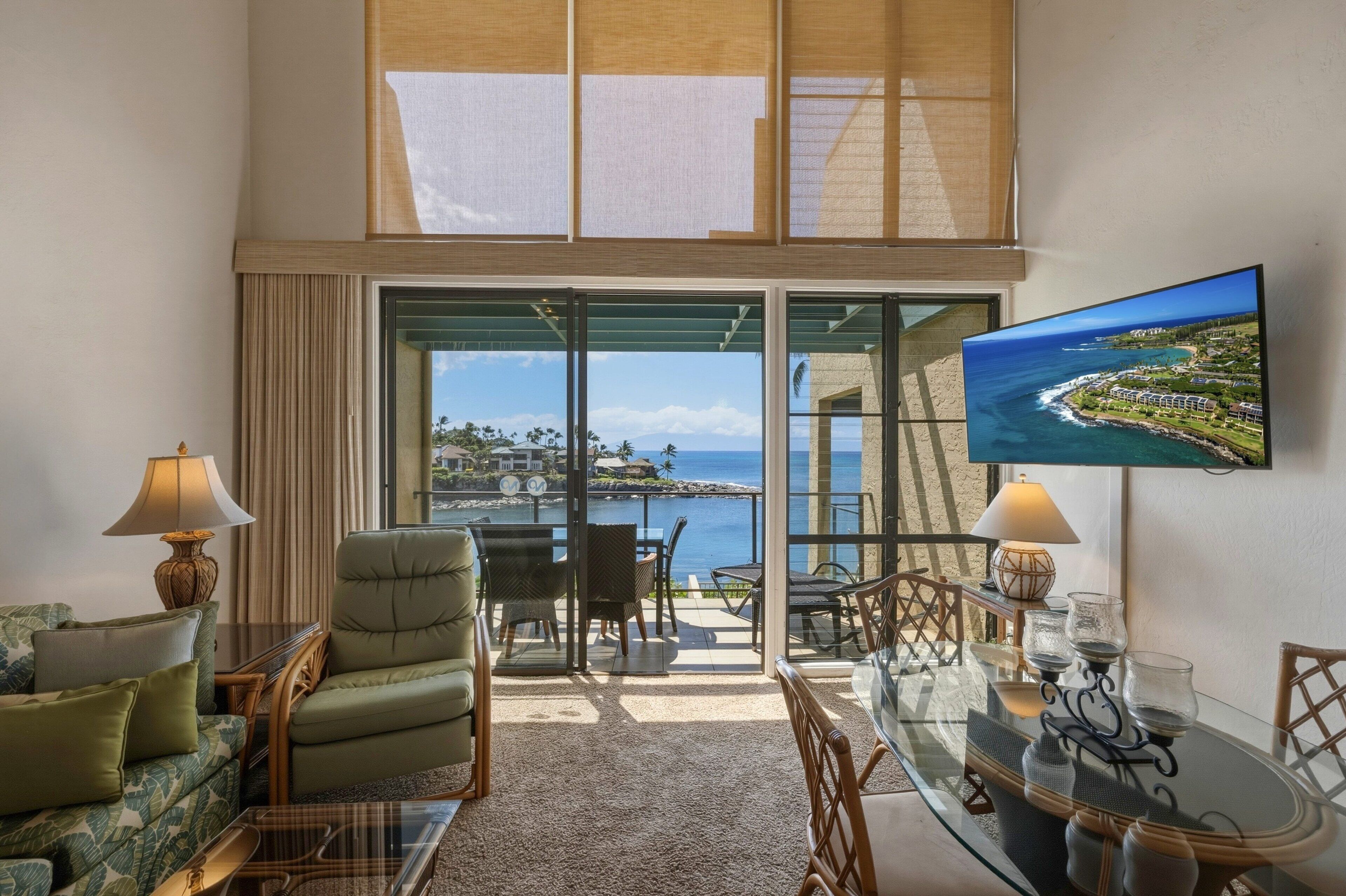 Een kamer met een bank, fauteuil, eettafel en stoelen, met een groot raam met uitzicht op zee.