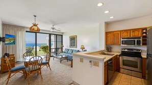 Eine moderne Küche mit Edelstahlgeräten, eine Essecke mit Glastisch und Stühlen sowie ein Wohnbereich mit Sofa und Blick auf den Ozean.