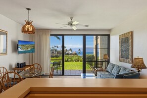 A room with a TV, a ceiling fan, a dining area with chairs, a sofa, and a view of the ocean through large sliding doors. - Napili Point Maui Oceanfront Stay 1br - Unit B25 1 Bedroom Condo (Lahaina)