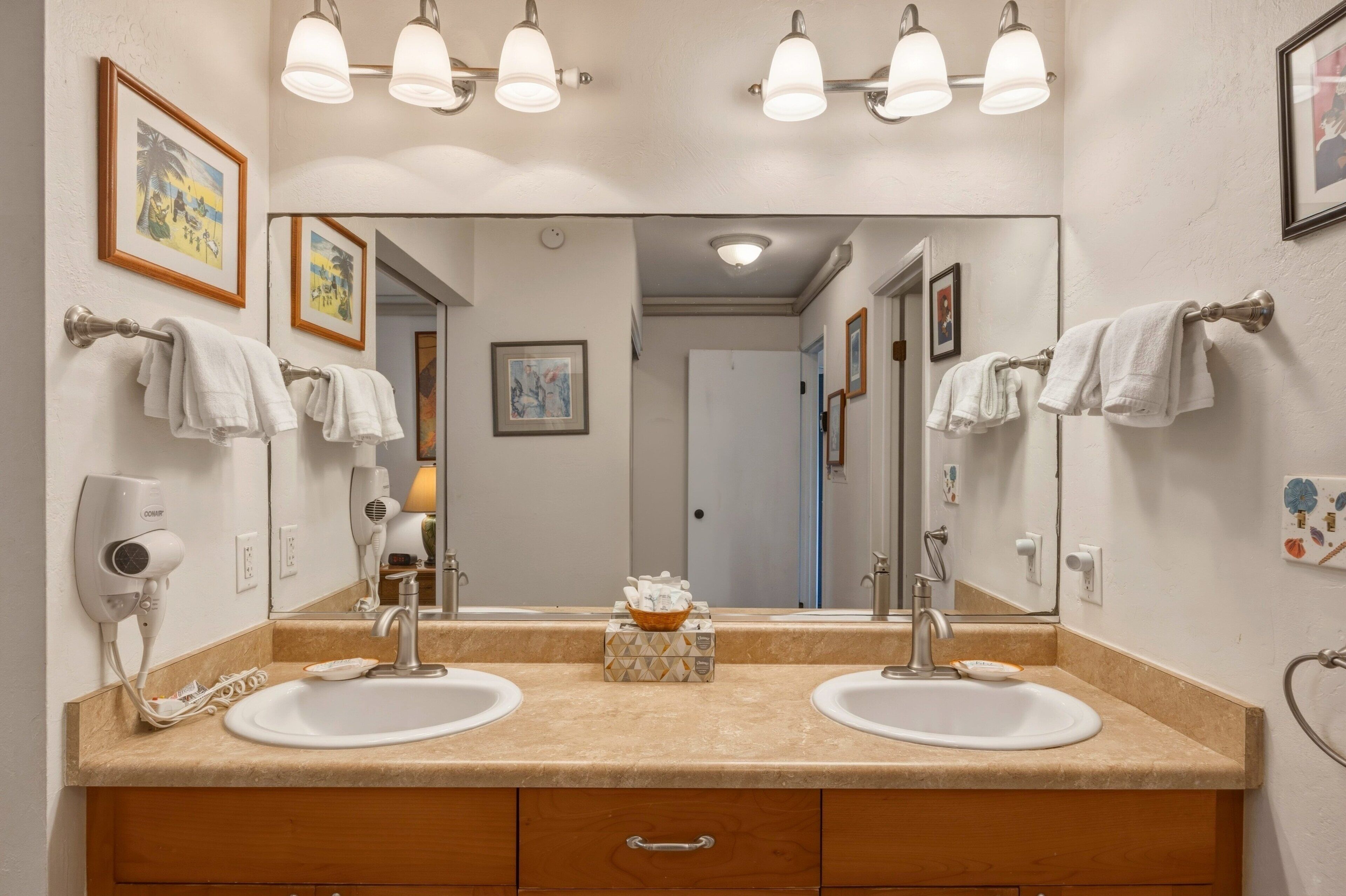 A bathroom with two sinks, a large mirror, towel racks, a hairdryer, and a small basket on the counter.
