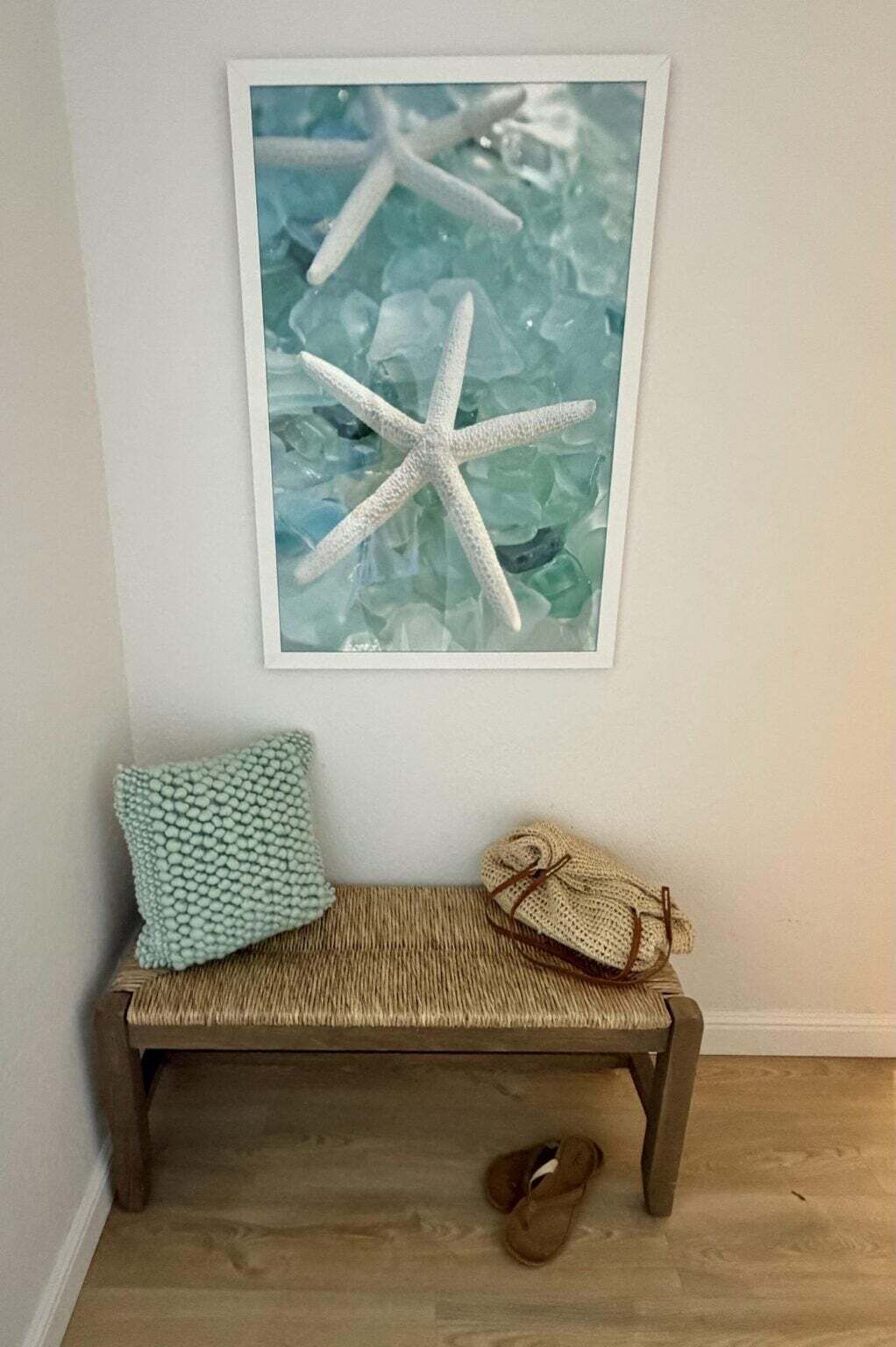 Una obra d'art emmarcada amb una estrella de mar a la paret, un banc de vímet amb coixins i un parell d'esliçons al sòl.