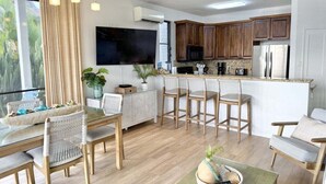 A modern kitchen with wooden cabinets, a large TV, and a dining area with wicker chairs. - Oceanfront Family Friendly 2br Maui - Unit B29 1 Bedroom Condo (Lahaina)