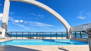 A swimming pool with a clear view of the sea, a tiled deck, and a large arched structure.