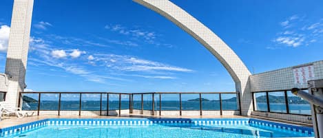 A swimming pool with a clear view of the sea, a tiled deck, and a large arched structure.