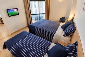 A hotel room with two beds, a TV on the wall, and a window with a view of buildings. - Clube XV (Santos)
