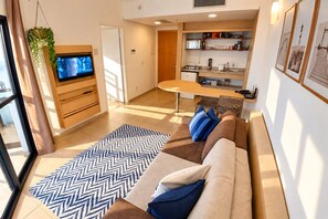 A modern living room with a sofa, a television, and a kitchen area with shelves. - Clube XV (Santos)